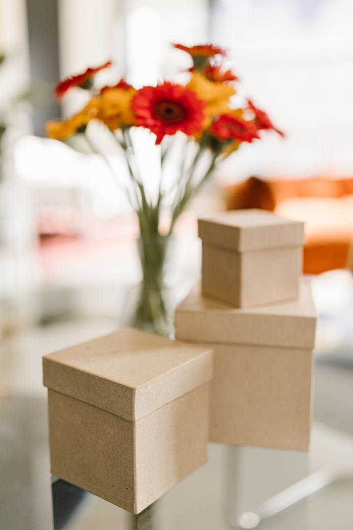 Minimalist shot of kraft boxes on a glass table with vibrant flowers in the background.